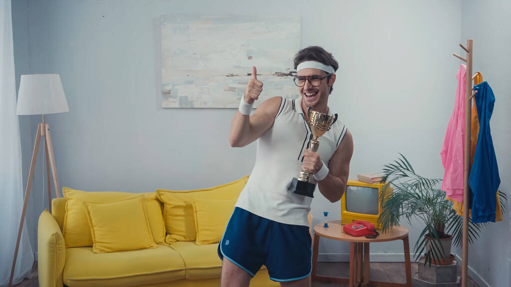 веселий спортсмен в окулярах тримає золотий трофей і показує великий палець вдома
 - Фото, зображення