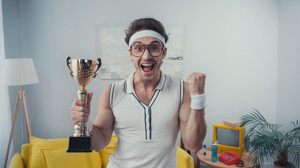 verbaasd winnaar in sportkleding en brillen met gouden trofee thuis - Foto, afbeelding
