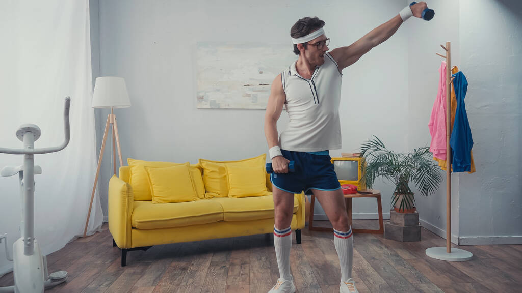 vicces sportoló szemüvegben gyakorló súlyzókkal a nappaliban, retro sport koncepció - Fotó, kép