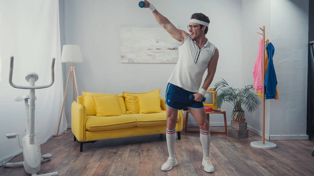positive sportsman lifting dumbbells and dancing in living room, retro sport concept - Photo, Image
