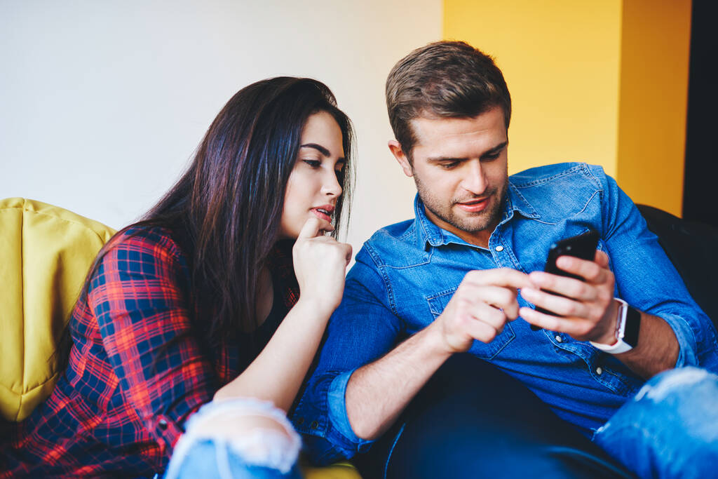 Young caucasian woman and man millennial choosing smth in booking app on mobile phone talking and discussing, couple in love looking social networks news via smartphone having good conversation - Photo, Image