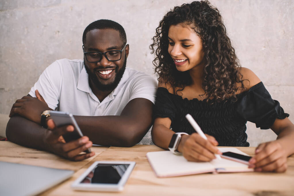 Millennial African American male and female friends laughing from received web content sitting at table and rejoicing during time for together learning, cheerful hipster guys using smartphone - Photo, Image