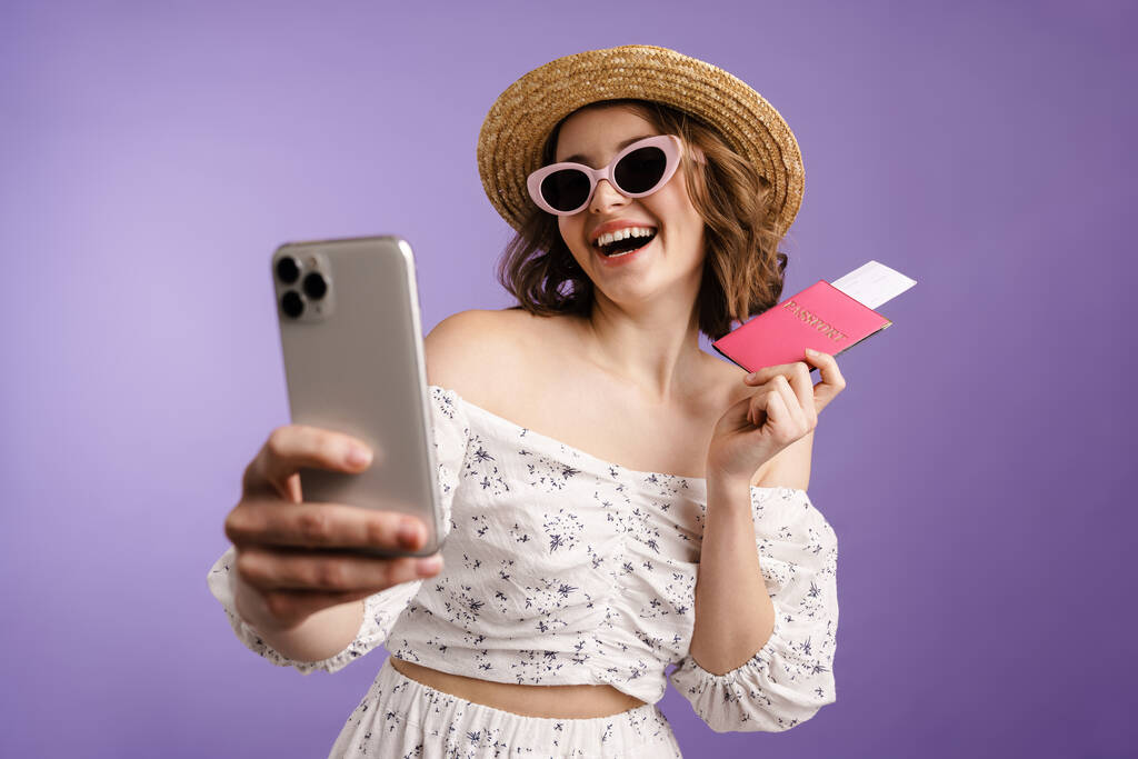 Mosolygó nő kezében útlevél jegyek, miközben szelfi a mobiltelefon elszigetelt felett lila fal - Fotó, kép