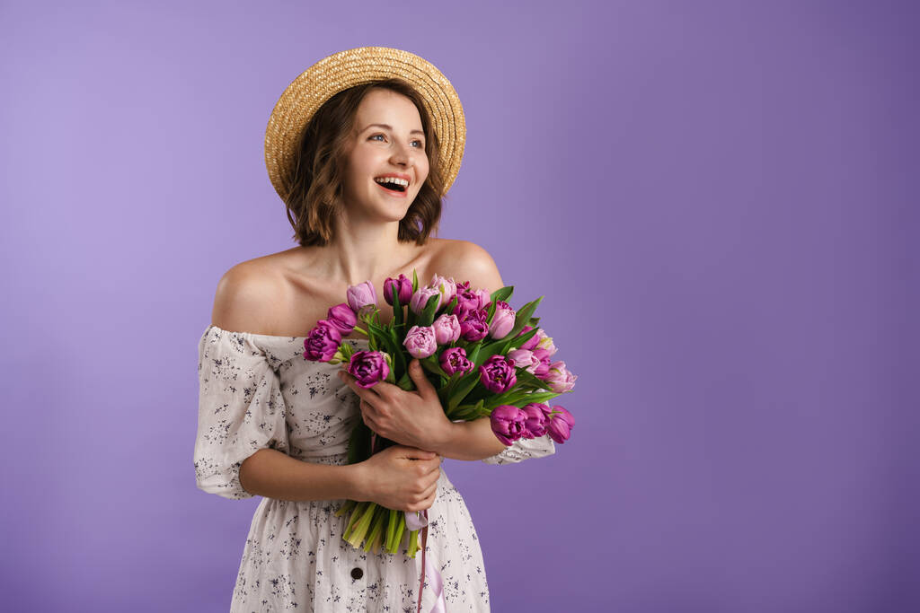 Fehér fiatal nő kalapban mosolyog, miközben pózol tulipán elszigetelt felett lila fal - Fotó, kép