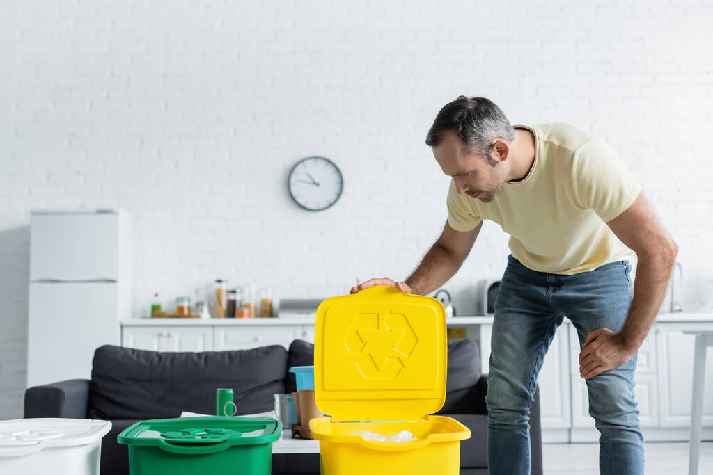 Homem olhando para lata de lixo com sinal de reciclagem na cozinha  - Foto, Imagem