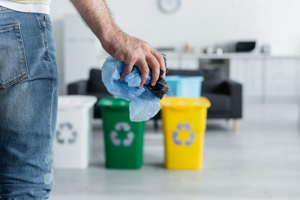 Ausgeschnittene Ansicht eines Mannes mit Plastiktüten in der Nähe verschwommener Dosen mit Recyclingschild zu Hause  - Foto, Bild