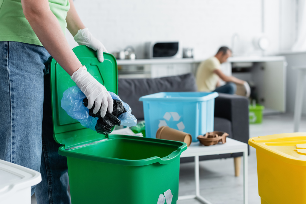 Lastik eldivenli bir kadının geri dönüşüm tabelasıyla plastik poşetleri çöp kutusuna koyması.  - Fotoğraf, Görsel