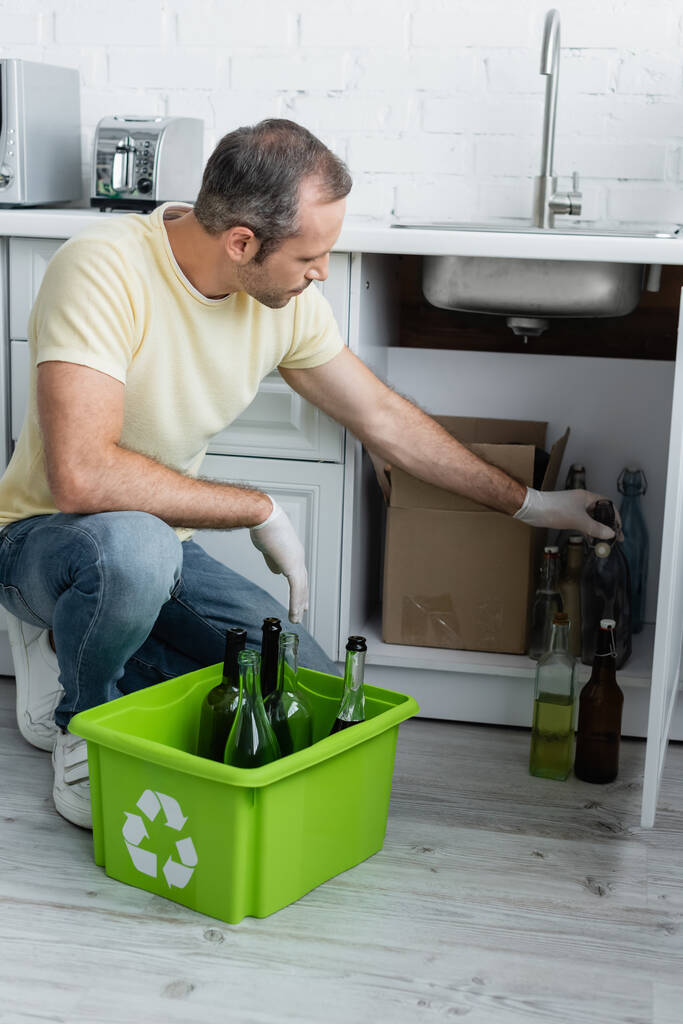 Homem tomando garrafa perto da caixa com sinal de reciclagem na cozinha  - Foto, Imagem