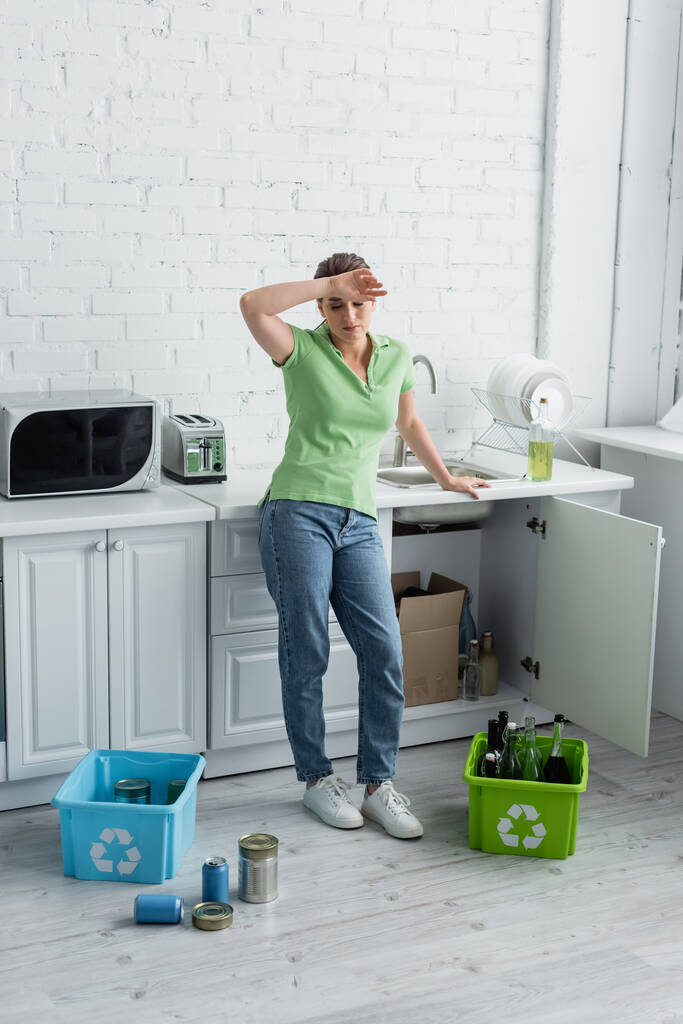 Femme fatiguée regardant des canettes près de la boîte avec panneau de recyclage dans la cuisine  - Photo, image
