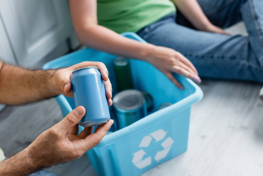 Vista recortada del hombre sosteniendo lata cerca de caja borrosa con signo de reciclaje y esposa en casa - Foto, imagen