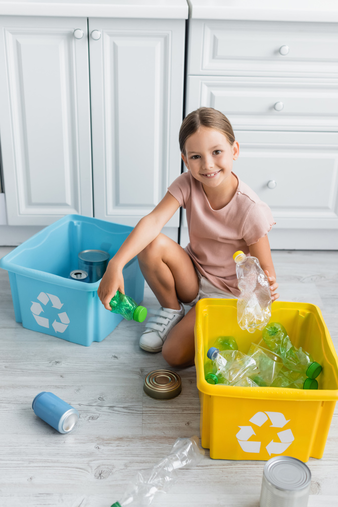 Muchacha sonriente sosteniendo botellas cerca de cajas con cartel de reciclaje en la cocina  - Foto, imagen