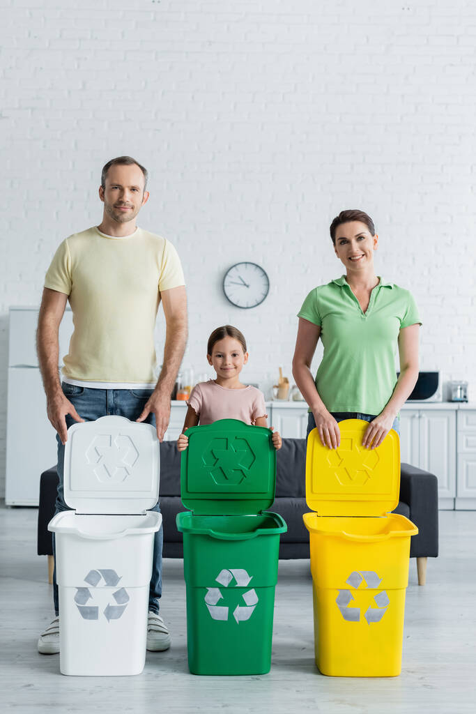 Positive Familie mit Tochter steht neben Mülltonnen mit Recyclingschild in Küche  - Foto, Bild