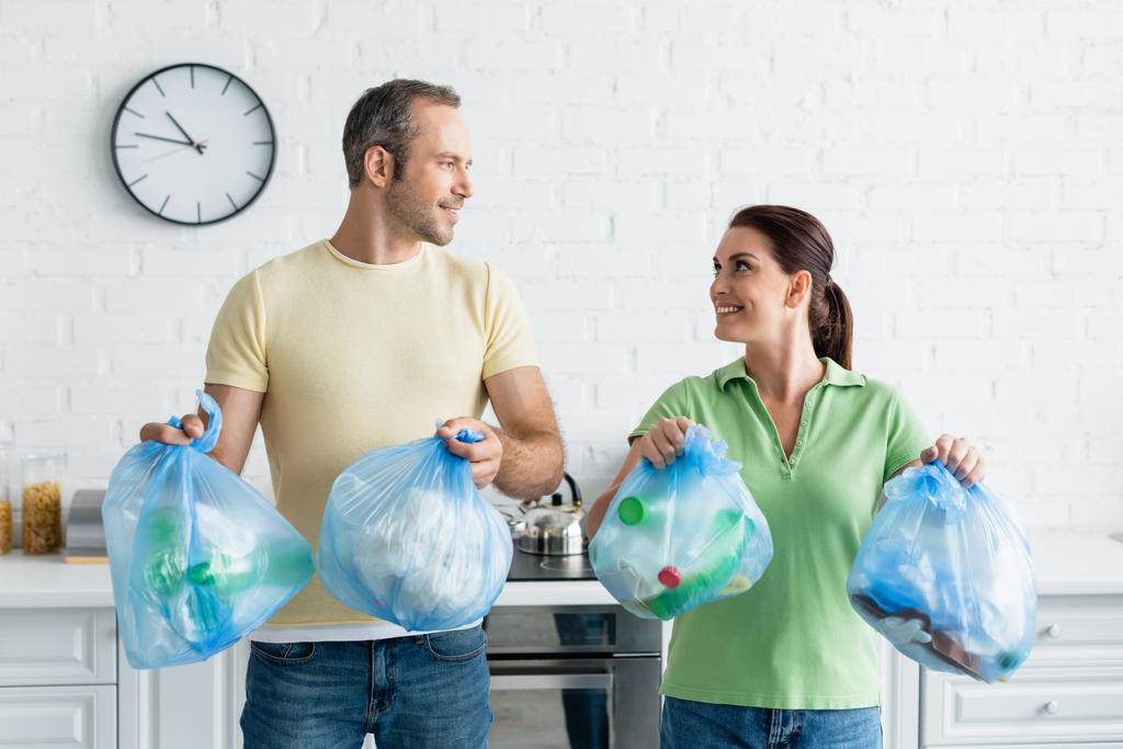 Couple souriant se regardant tout en tenant des sacs poubelle dans la cuisine  - Photo, image