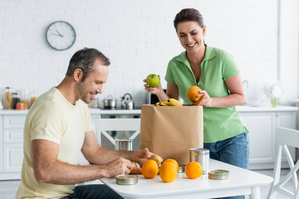 Mann hält Essen neben fröhlicher Frau und Papiertüte in Küche  - Foto, Bild