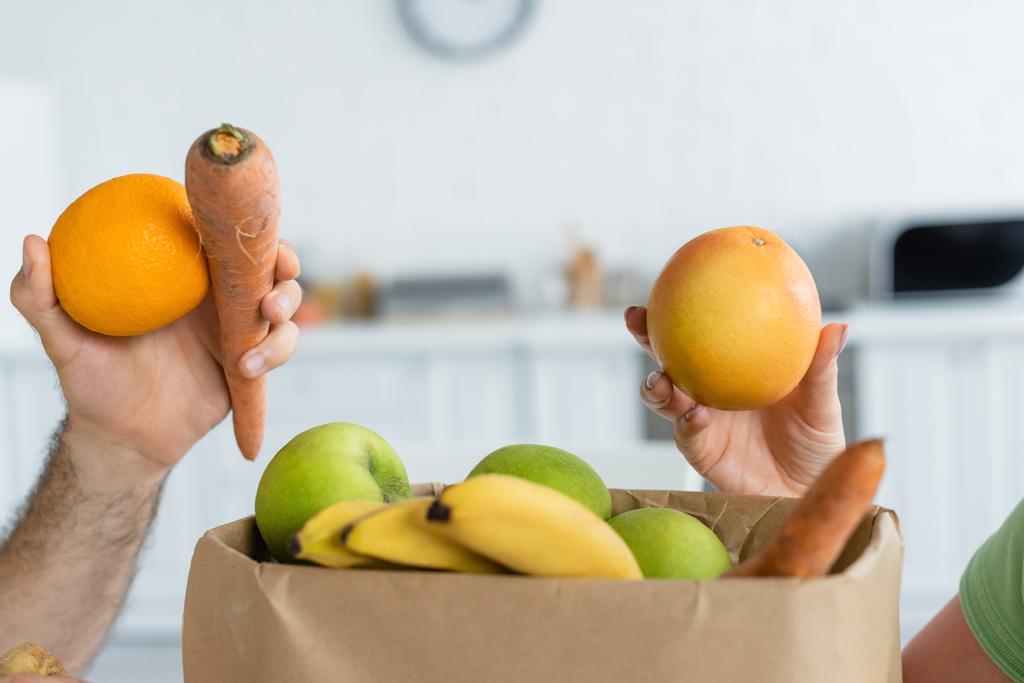 Обрезанный вид на пару, держащую фрукты возле бумажного пакета на кухне  - Фото, изображение