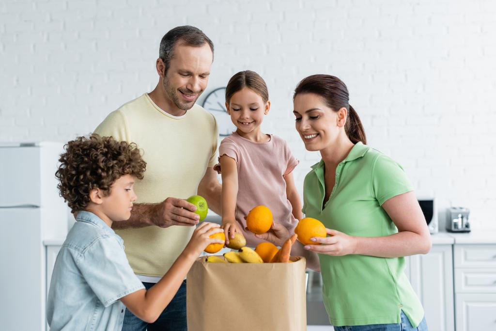 Famille joyeuse avec des enfants tenant des fruits près du sac en papier dans la cuisine  - Photo, image