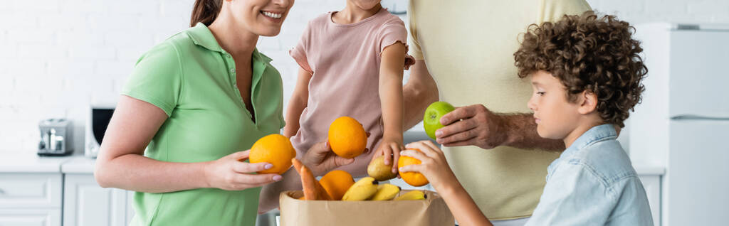 Mujer sonriente sosteniendo fruta cerca de la familia y bolsa de papel en la cocina, pancarta  - Foto, Imagen