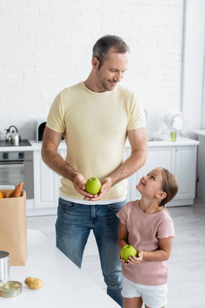 Sonriente padre e hija sosteniendo manzanas cerca de la bolsa de papel en la cocina  - Foto, Imagen