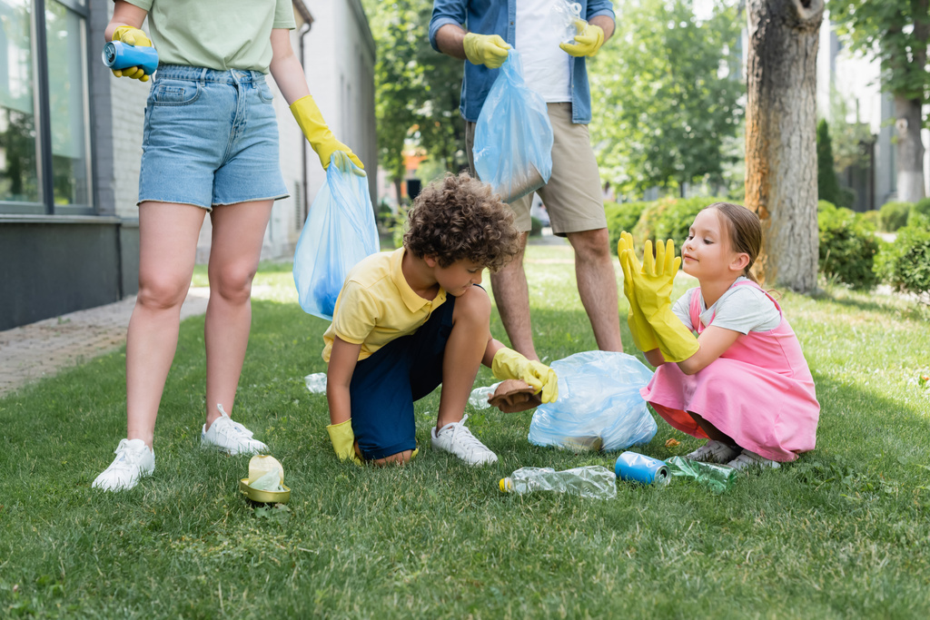 Kinder in Gummihandschuhen auf Rasen neben Eltern mit Müllsäcken  - Foto, Bild