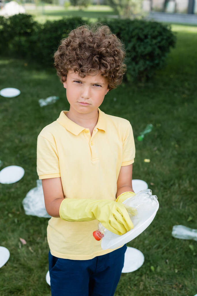 Хлопчик в гумових рукавичках тримає сміття і дивиться на камеру на відкритому повітрі
  - Фото, зображення