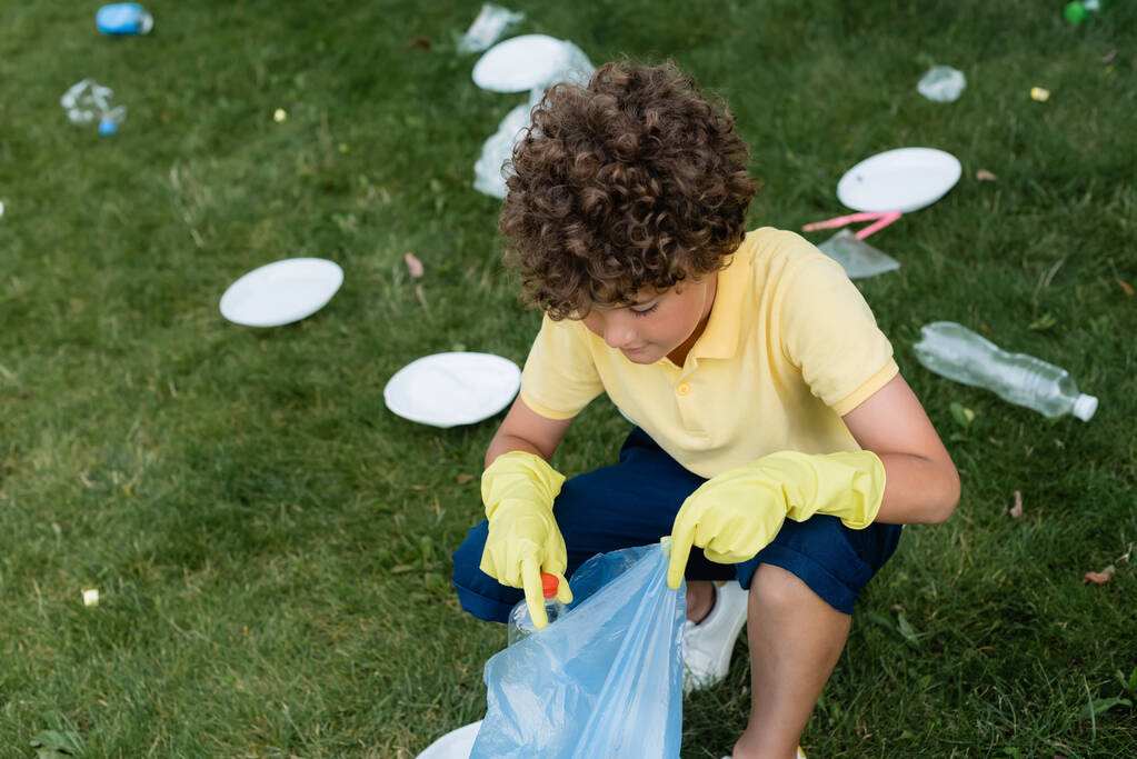 Hoge hoek uitzicht van jongen in rubber handschoenen verzamelen van afval in zak op gazon  - Foto, afbeelding
