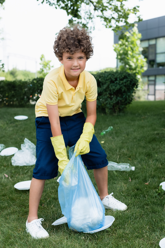 Positieve jongen in rubber handschoenen houden tas in de buurt van afval op gras  - Foto, afbeelding