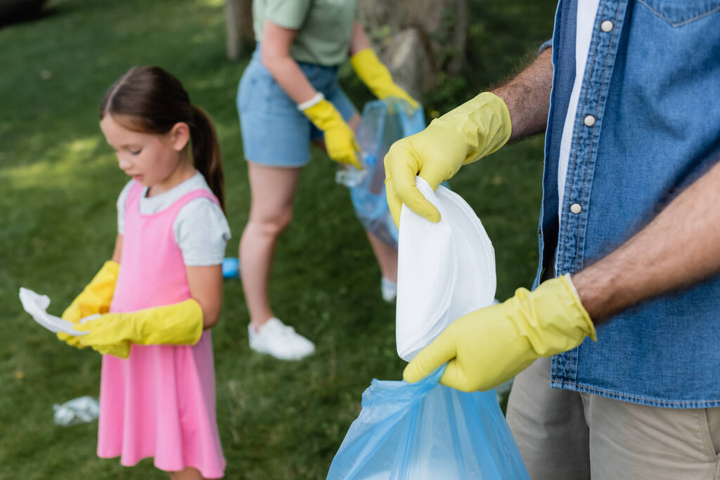 Man in rubber gloves putting trash in bag near blurred daughter and wife outdoors  - Photo, Image