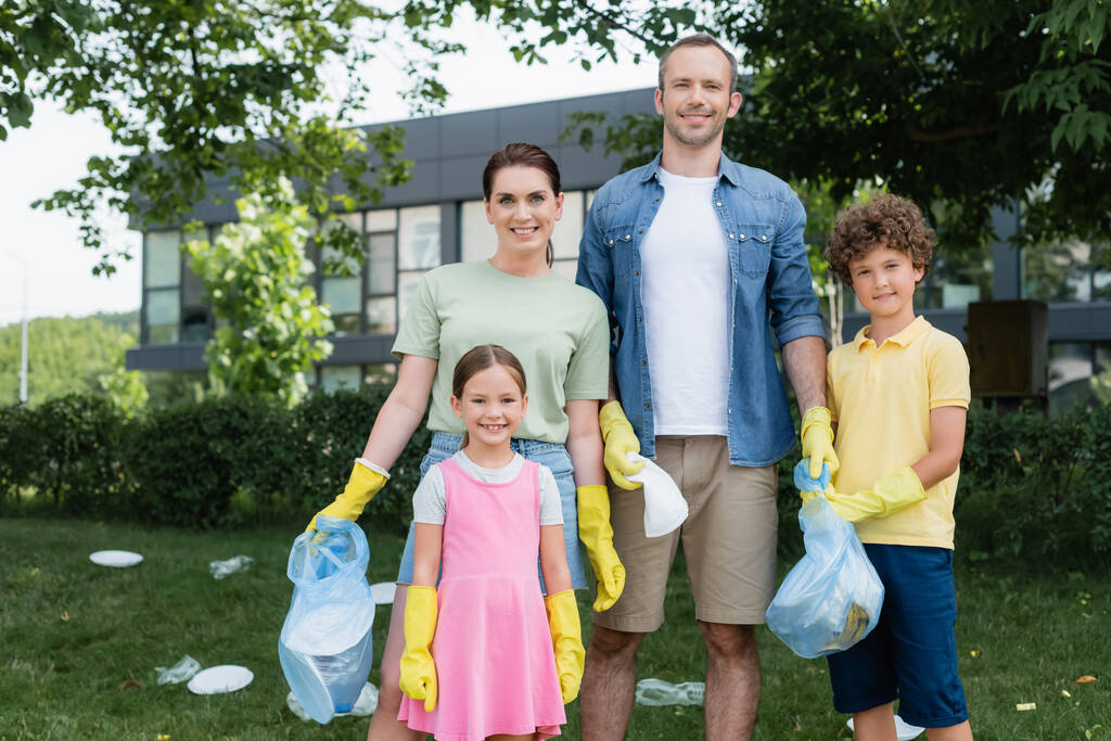 Pozytywna rodzina z dziećmi w gumowych rękawiczkach trzymająca worki na śmieci na zewnątrz  - Zdjęcie, obraz