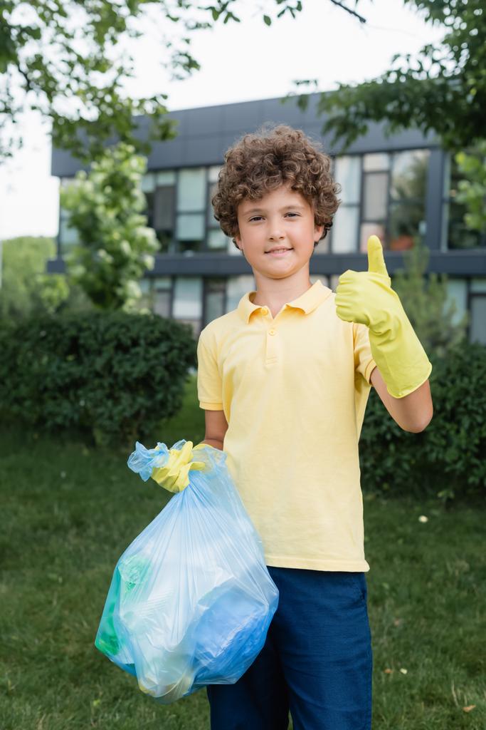 Lächelnder Junge in Gummihandschuhen hält Müllsack in der Hand und zeigt wie eine Geste im Freien  - Foto, Bild