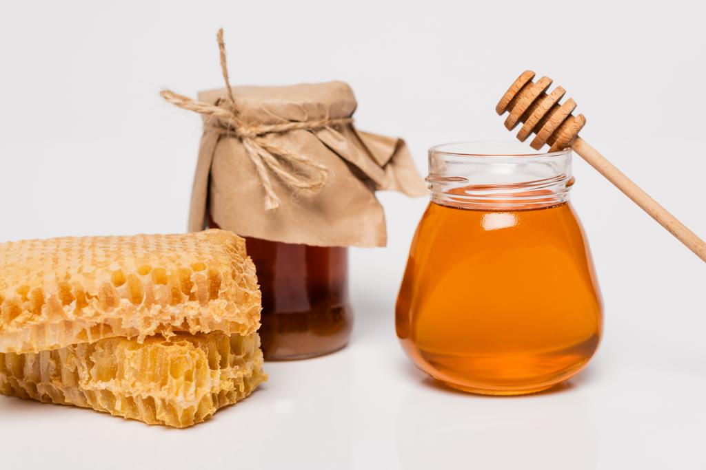 jarros com mel amarelo e marrom perto de favo de mel e mergulhador de madeira no branco - Foto, Imagem