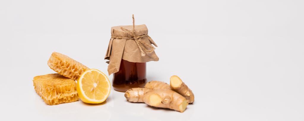 tarro de miel cubierto con papel artesanal cerca de limón fresco, raíz de jengibre y panal en blanco, bandera - Foto, Imagen