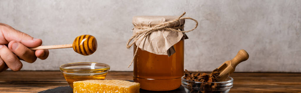 vista cortada da mão masculina com mergulhador de madeira perto favo de mel, jarra e tigelas com mel e sementes de anis em fundo cinza, banner - Foto, Imagem