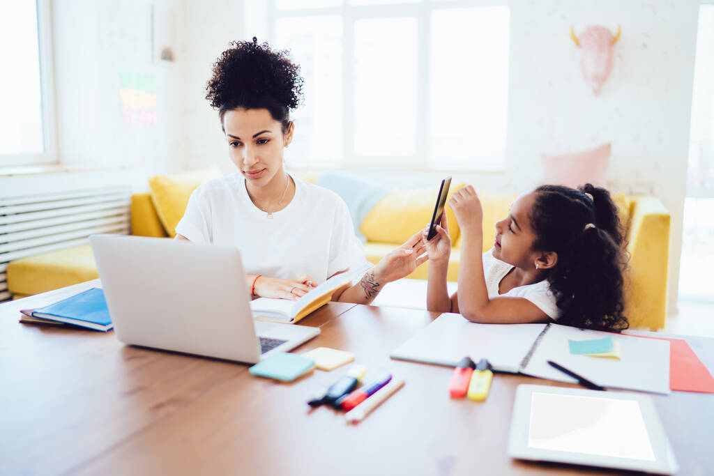 Ethnische Mädchen lenkt Mutter davon ab, Hausaufgaben im Notizblock mit dem Handy zu überprüfen und Fotos zu machen, wie sie zusammen am Tisch im hellen Raum sitzen - Foto, Bild