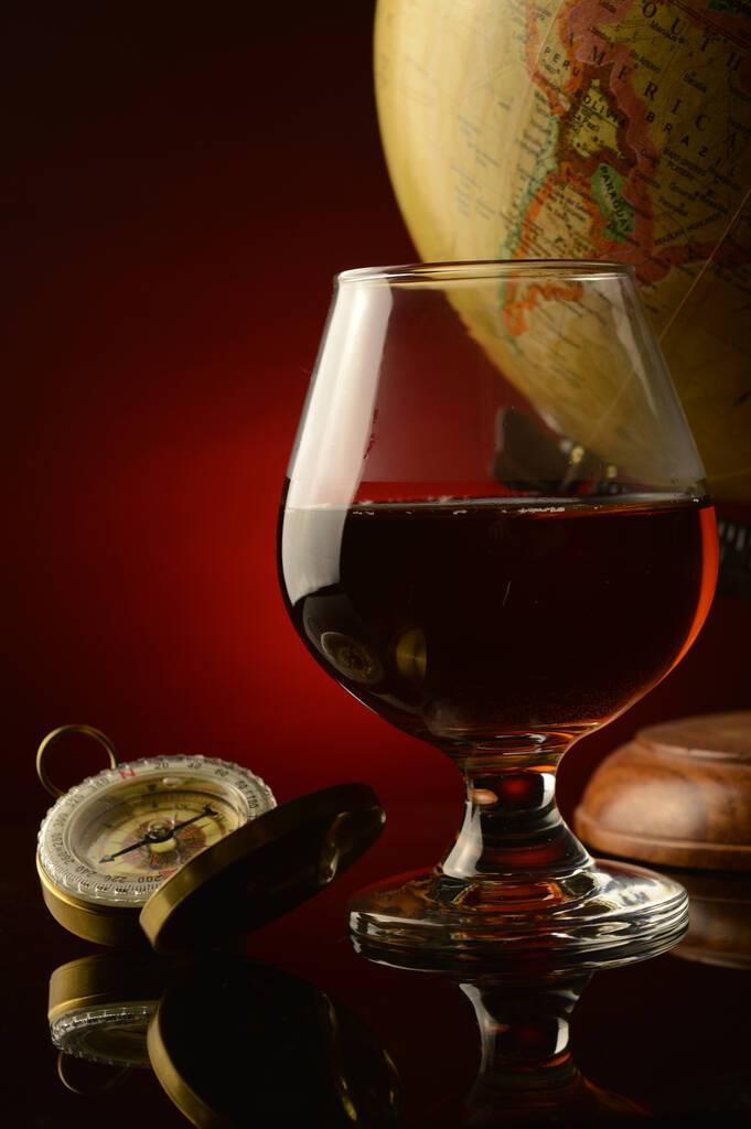 Une faible profondeur de champ se concentre sur un verre de Cognac avec une boussole et un globe antique avec un fond rouge et noir réfléchissant foncé. - Photo, image