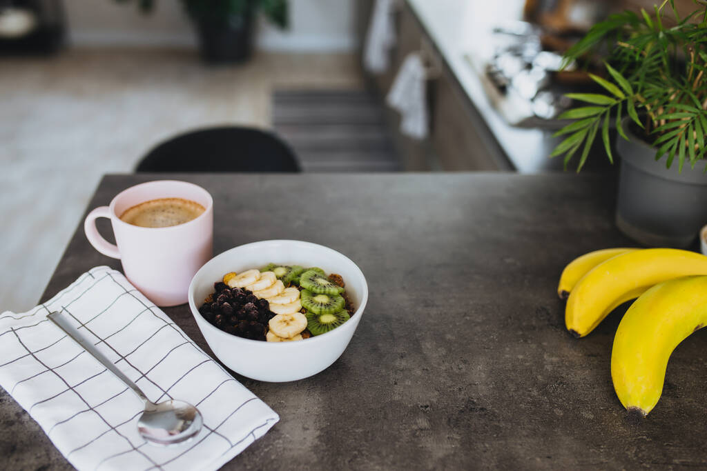 Taza de café rosa, tazón con kiwi de frutas tropicales picadas y plátano, arándanos, cuchara en el mostrador de bar en la elegante cocina loft. Fondo borroso. Foto de alta calidad - Foto, imagen