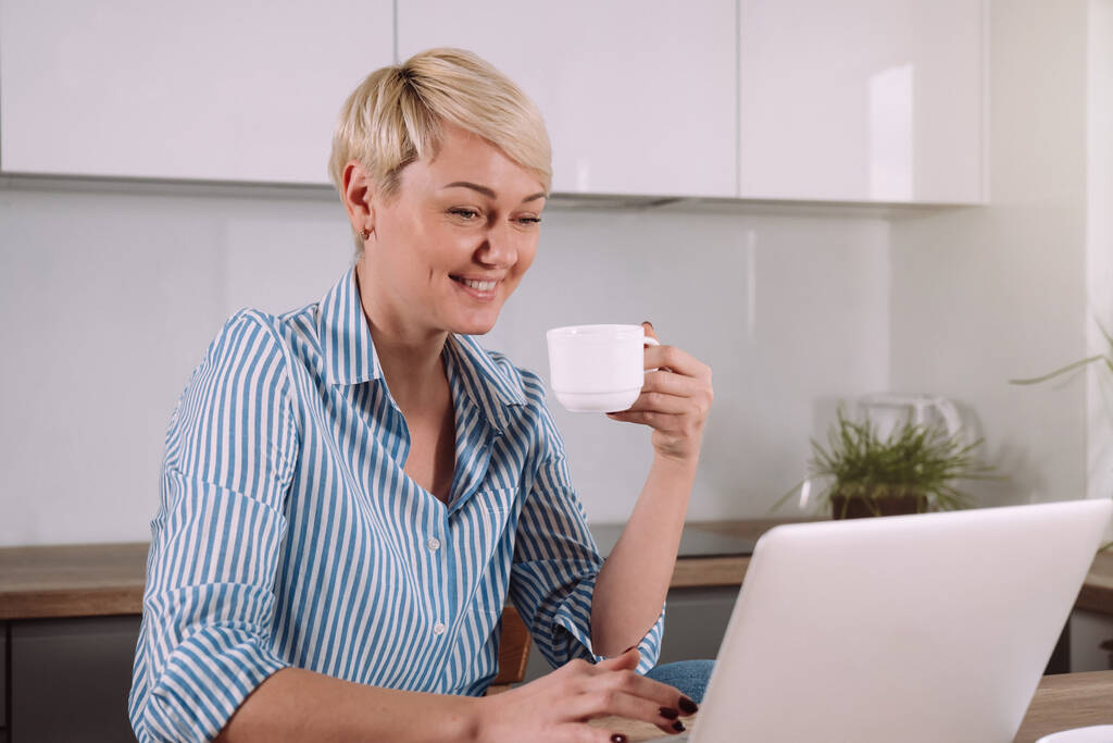 陽気な若い女性がコーヒーを飲みながら、自宅でキッチンに座っている間、ラップトップコンピュータ上のビデオ会議で話しています - 写真・画像
