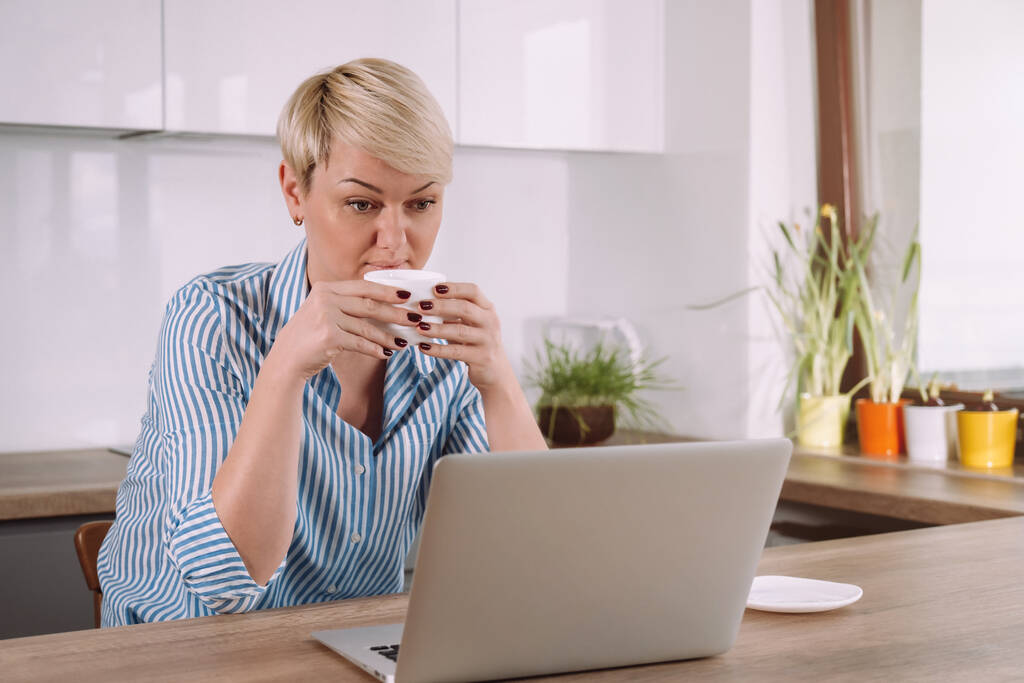 Freelancer donna con tazza di caffè e parlando in videoconferenza sul computer portatile mentre seduto in cucina a casa - Foto, immagini