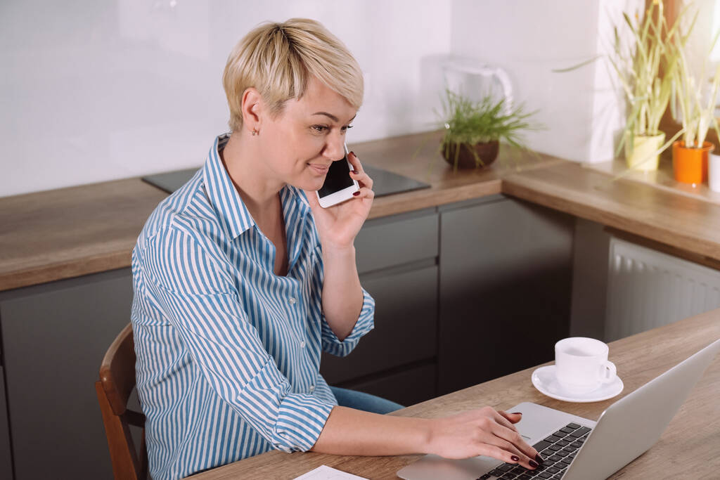 Женщина, работающая дома на ноутбуке, разговаривает по телефону и делает заметки с помощью портативного компьютера на кухне - Фото, изображение