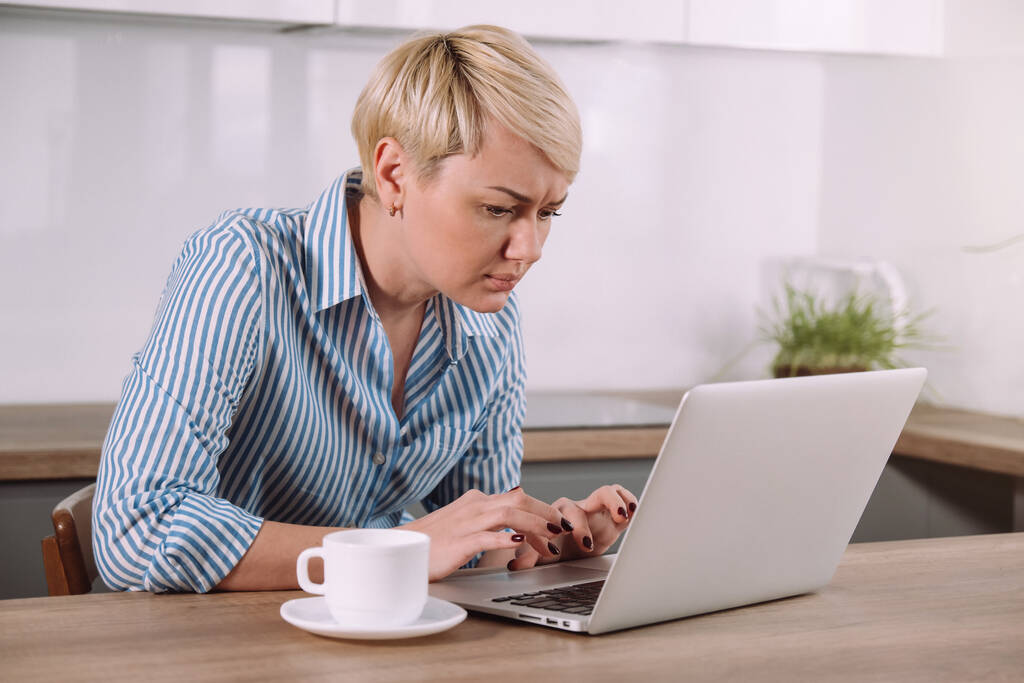 Фокусована серйозна жінка, що працює на ноутбуці вдома Жінка використовує портативний комп'ютер на кухні
 - Фото, зображення
