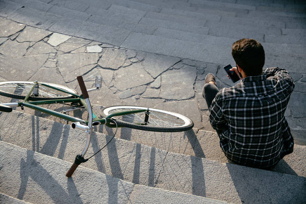 Top view on trendy man using a mobile phone, while resting on steps, sitting near the bike outdoors. View from back. - Photo, Image