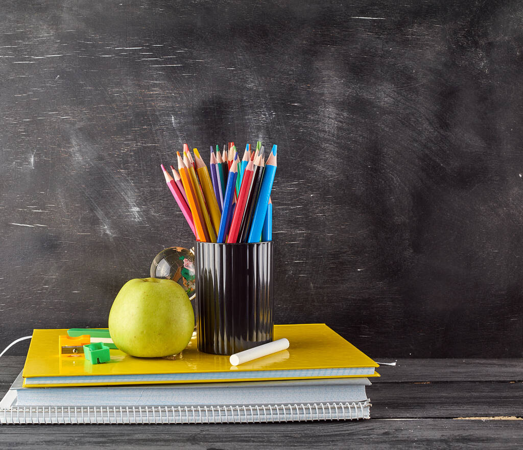 dizüstü bir yığın üzerinde olgun yeşil elma, boş bir siyah tebeşir tahtası arka planda çok renkli ahşap kalemler ile siyah kırtasiye cam, okul kavramıgeri - Fotoğraf, Görsel