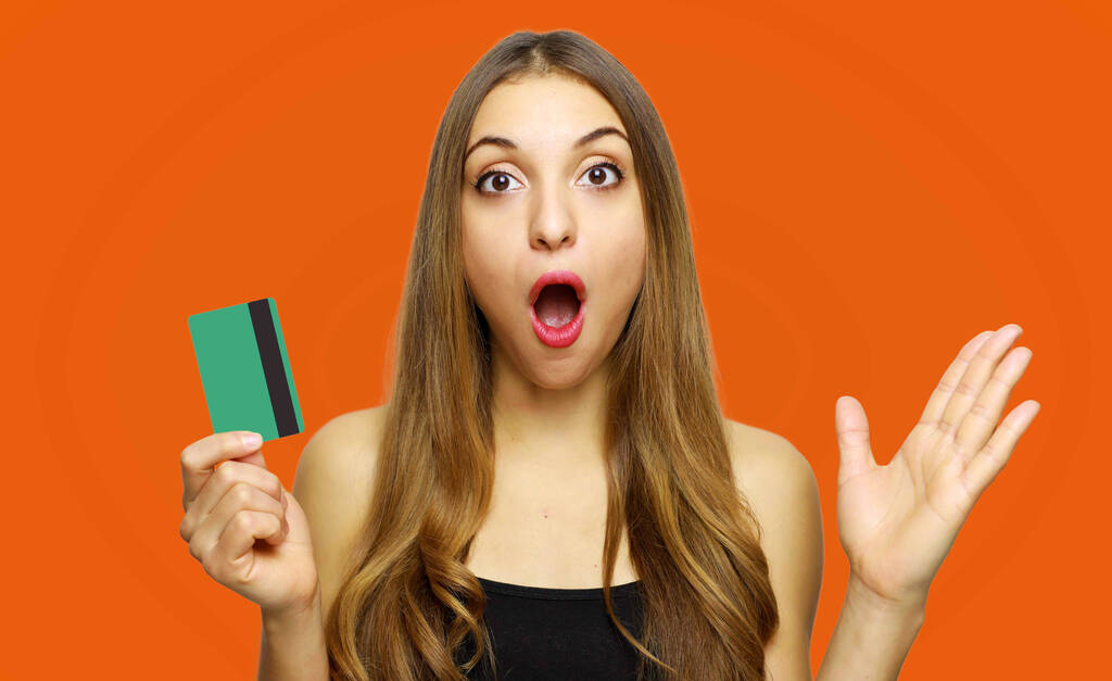 Sorprendido emocionado mujer joven sorprendida sosteniendo tarjeta de crédito sobre fondo naranja - Foto, Imagen