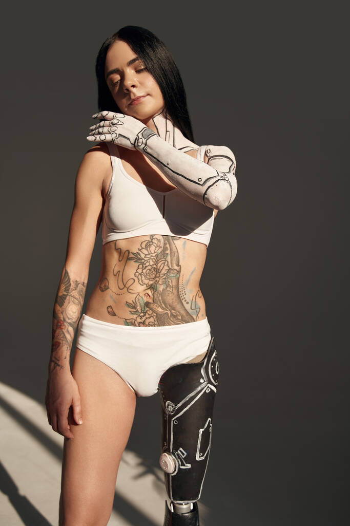 Νεαρή γυναίκα με τατουάζ, με ρομπότ ή σάιμποργκ, που φοράει εσώρουχα, κοιτάζει το χέρι της και ποζάρει στην κάμερα. Στιγμιότυπο. Στοκ φωτογραφία - Φωτογραφία, εικόνα