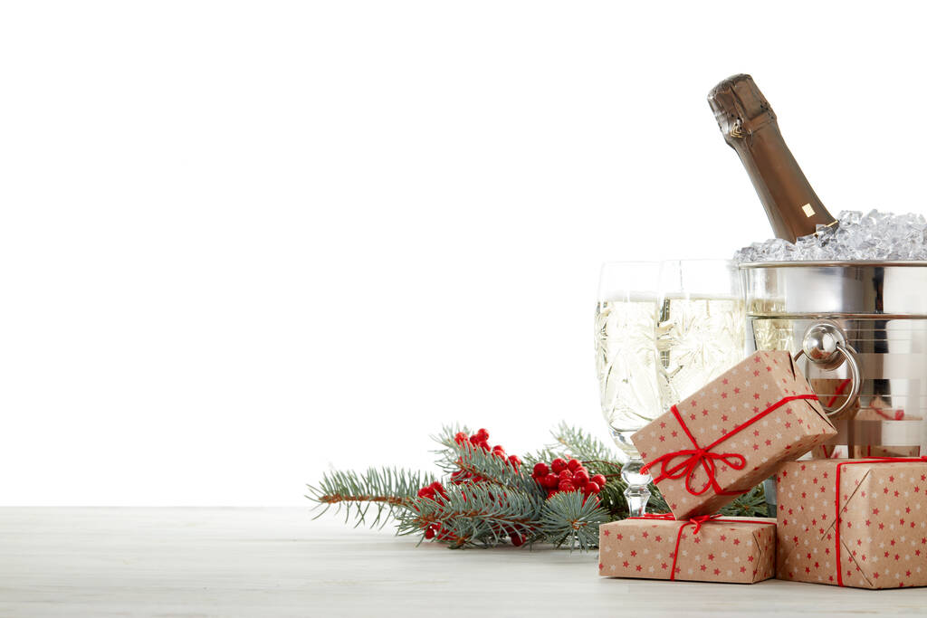 Zátiší s lahví šampaňského stojící v kbelíku s ledem, šampaňskými flétnami, dárkovými krabicemi a jedlovou větví na dřevěném stole na bílém pozadí s kopírovacím prostorem. Vánoce a Nový rok. Pozdrav - Fotografie, Obrázek