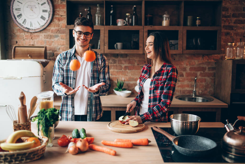 El joven marido hace malabares con naranjas en la cocina mientras su esposa cocina el desayuno de frutas. Hombre y mujer preparando ensalada de verduras, familia feliz juntos - Foto, Imagen