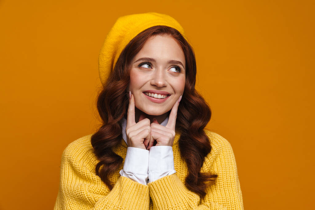 Mujer joven feliz con el pelo rojo largo en suéter y boina de pie sobre fondo amarillo de la pared, mirando a un lado - Foto, imagen
