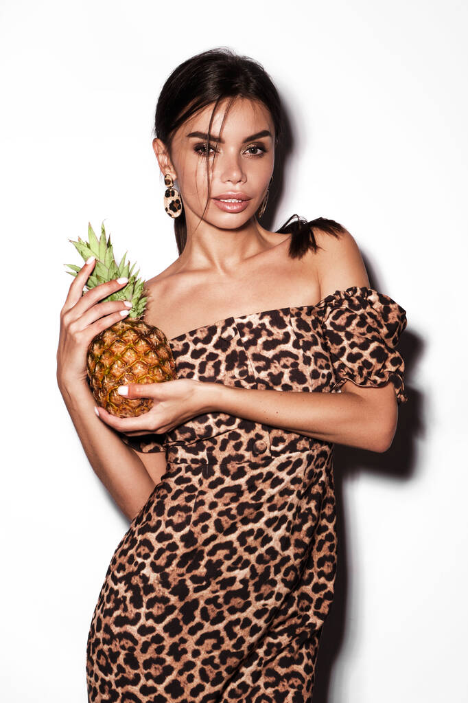 Schöne sexy Frau mit Ananas in den Händen in einem Leopardenkleid und Ohrringen, mit klassischem Smokey Make-up und einer modischen Frisur. Schönheitsgesicht. - Foto, Bild