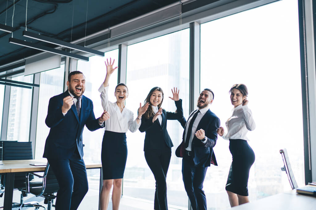 Grupo alegre de gente de negocios con las manos levantadas de pie sonriendo celebrando el éxito y el buen trabajo en equipo en el edificio de oficinas moderno - Foto, imagen