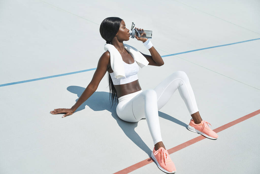 Якась вродлива африканка у спортивному одязі п "є воду, сидячи на відкритому повітрі. - Фото, зображення