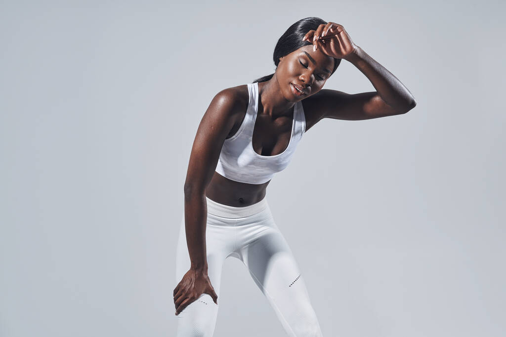 灰色の背景を背景に立っている間、額に触れるスポーツ服の若いアフリカの女性の疲れ - 写真・画像